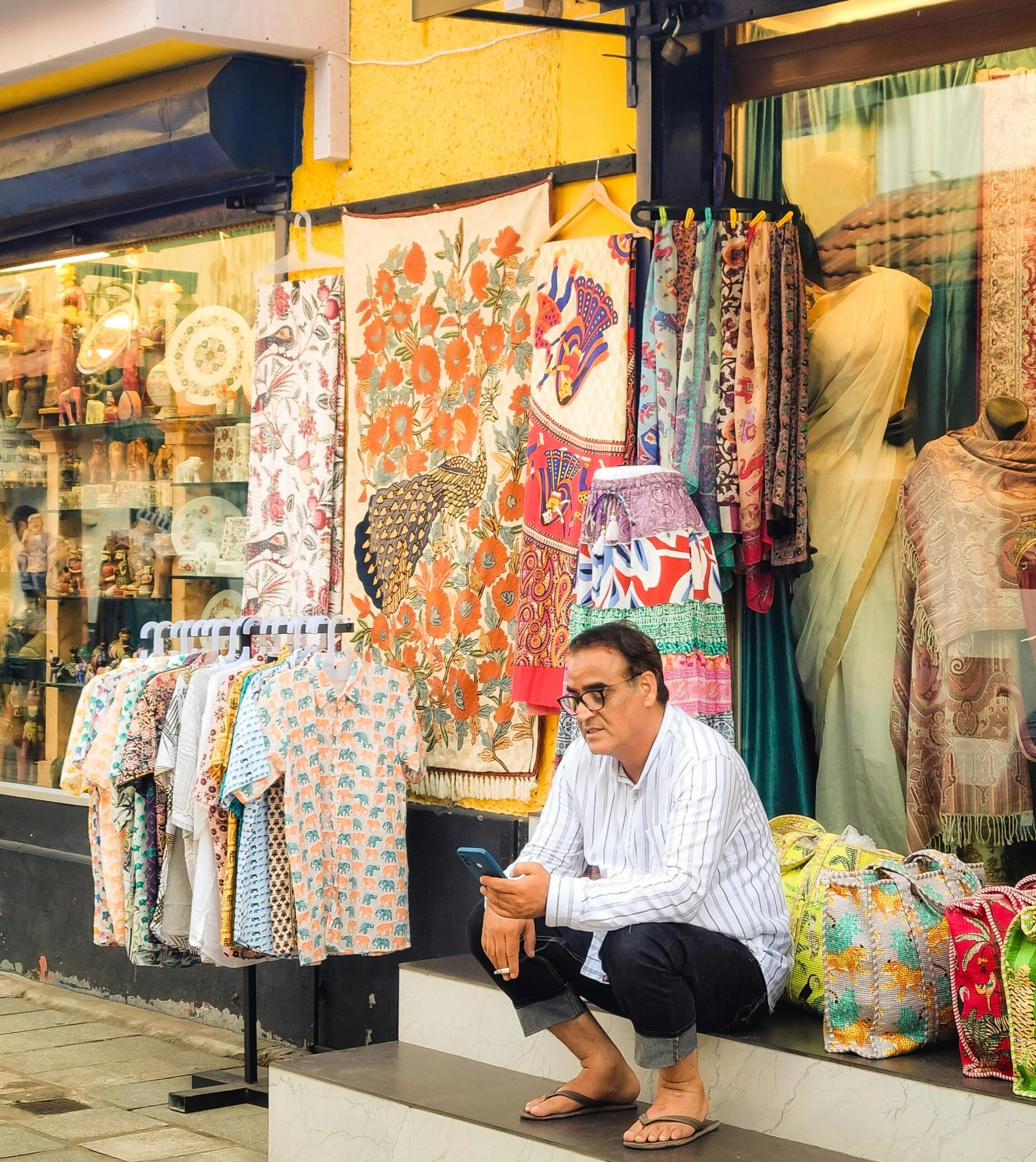 A local vendor sits outside a vibrant textile shop, displaying colorful fabrics and clothing.